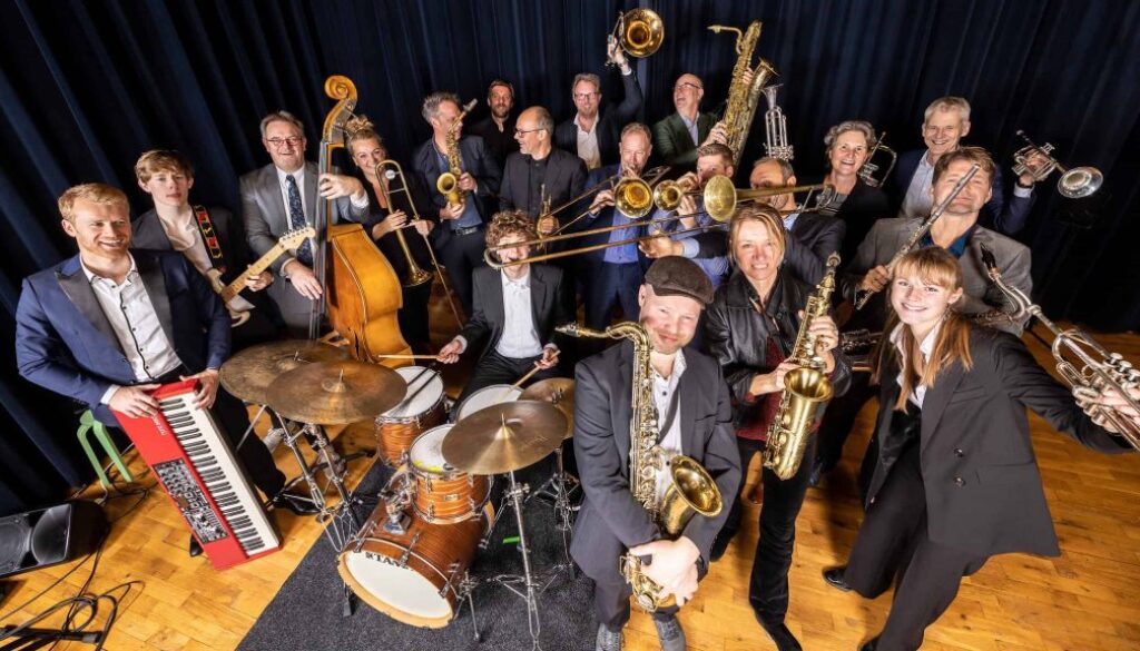 Een enthousiast orkest van jazz- en soulmusici met verschillende instrumenten, including gitaar, drums, saxofon, trompet en keyboard, op een podium met donkerblauw gordijn.