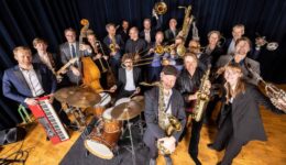 Een enthousiast orkest van jazz- en soulmusici met verschillende instrumenten, including gitaar, drums, saxofon, trompet en keyboard, op een podium met donkerblauw gordijn.