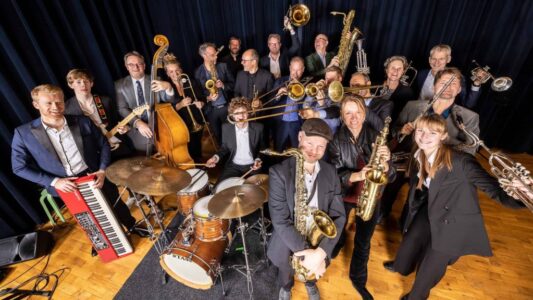 Een enthousiast orkest van jazz- en soulmusici met verschillende instrumenten, including gitaar, drums, saxofon, trompet en keyboard, op een podium met donkerblauw gordijn.