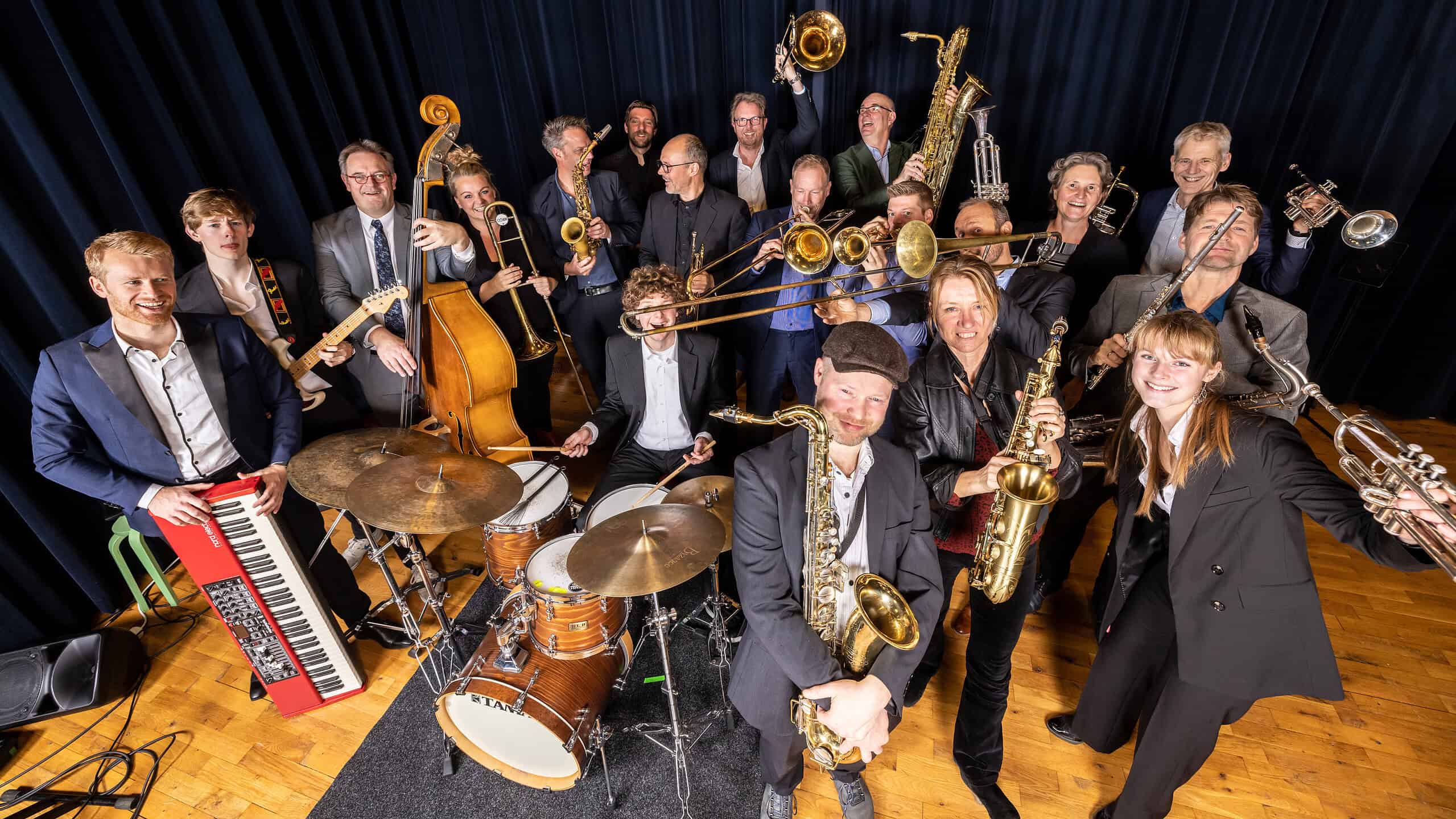 Een enthousiast orkest van jazz- en soulmusici met verschillende instrumenten, including gitaar, drums, saxofon, trompet en keyboard, op een podium met donkerblauw gordijn.