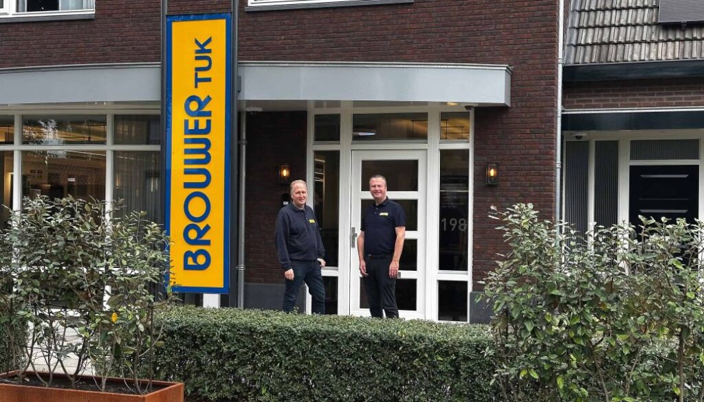 Bedrijfsgebouw van RTV SLOS met twee medewerkers voor de ingang, signaalbord met de naam, moderne gevel, personeel in uniforme kleding, lokale omroep, televisiestudio, radiostation, professionele uitstraling, communicatie, media-support, nieuwsvoorziening.