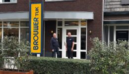 Bedrijfsgebouw van RTV SLOS met twee medewerkers voor de ingang, signaalbord met de naam, moderne gevel, personeel in uniforme kleding, lokale omroep, televisiestudio, radiostation, professionele uitstraling, communicatie, media-support, nieuwsvoorziening.