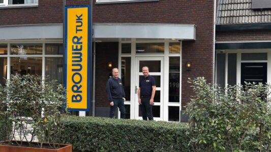 Bedrijfsgebouw van RTV SLOS met twee medewerkers voor de ingang, signaalbord met de naam, moderne gevel, personeel in uniforme kleding, lokale omroep, televisiestudio, radiostation, professionele uitstraling, communicatie, media-support, nieuwsvoorziening.