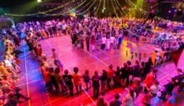 Dansfeest met kleurrijke verlichting en feestende mensen op een sporthalvloer, georganiseerd door RTV SLOS. Een gezellige evenementendoor waar iedereen danst en plezier heeft.