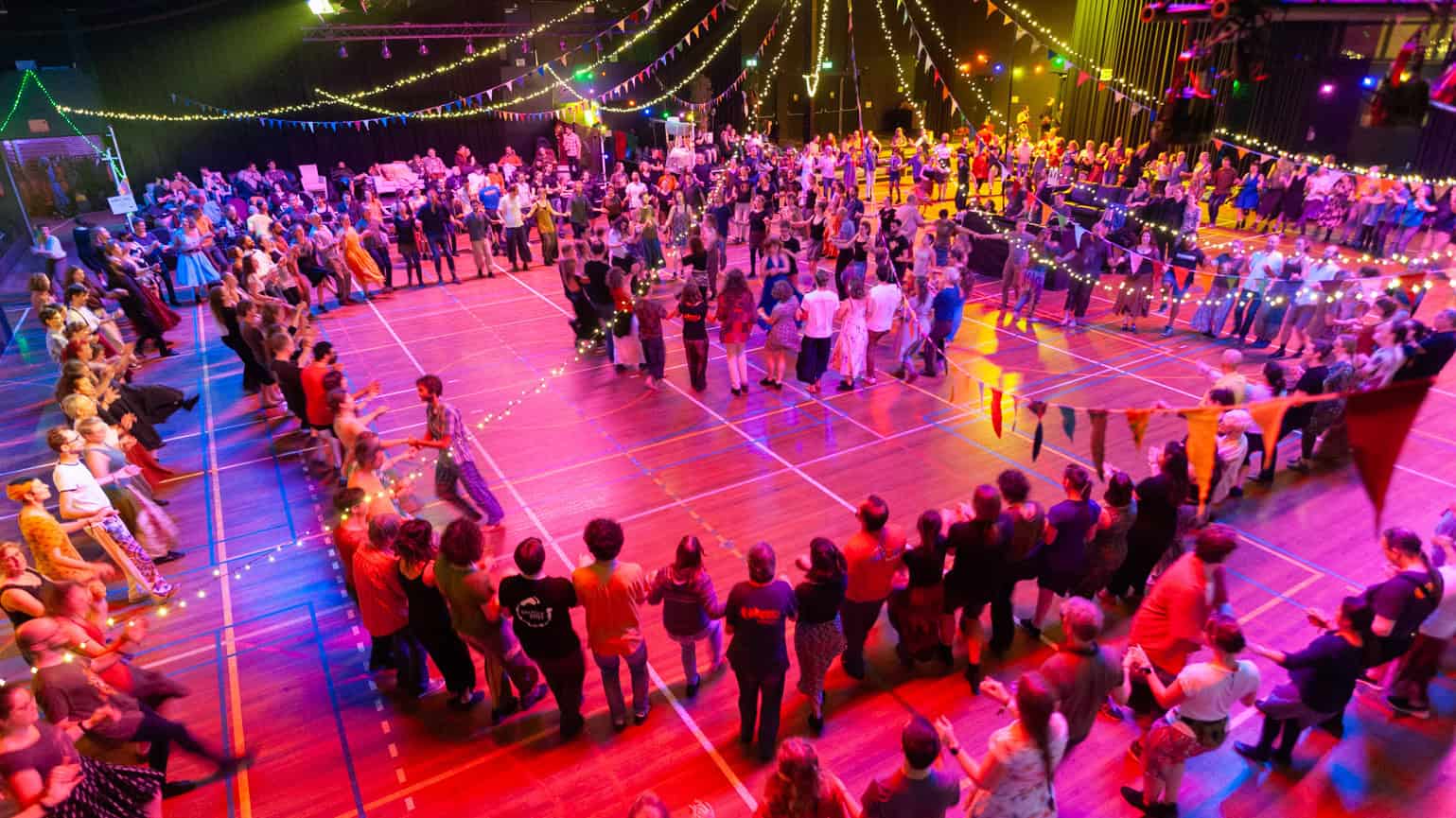 Dansfeest met kleurrijke verlichting en feestende mensen op een sporthalvloer, georganiseerd door RTV SLOS. Een gezellige evenementendoor waar iedereen danst en plezier heeft.