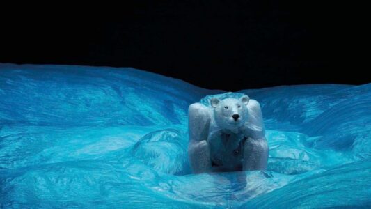 Robuuste ijsbeerfiguur op ijslandschap, gemaakt van kunstmatig ijs en plastic, afgebeeld bij nacht met helder blauw ijs en donkere lucht, symbool voor natuurbescherming en kunstprojecten.