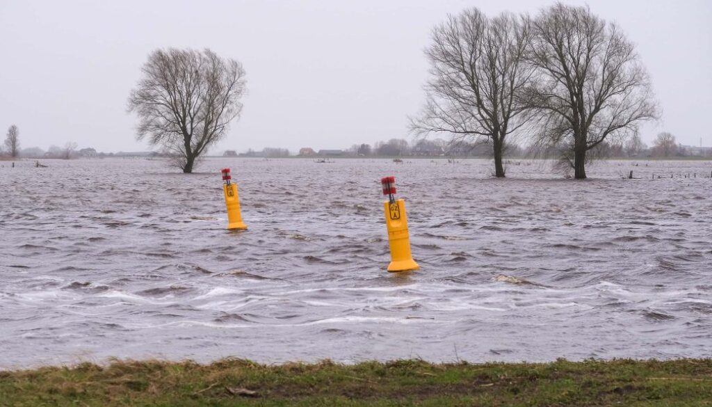 Overstromingsgebied in Nederland met waterniveau hoger dan normaal, waarschuwingsborden en blankstaande bomen. Noodweer en wateroverlast door extreme regenval en hoge waterstanden in de regio.
