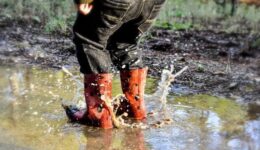 Kind met oranje rubberlaarzen waden door modderig water in een natuurgebied.