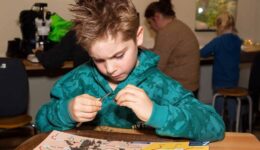 Meneer jeugdig man met speels gedrag en opvallend haar tijdens een kinderactiviteit bij RTV SLOS, bezig met een wetenschappelijke experiment of knutselproject op een houten tafel.
