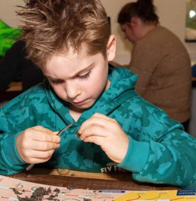 Meneer jeugdig man met speels gedrag en opvallend haar tijdens een kinderactiviteit bij RTV SLOS, bezig met een wetenschappelijke experiment of knutselproject op een houten tafel.