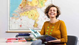 Gezellige vrouw leest kleurrijk kinderboek in klaslokaal met stapel kinderboeken op tafel, achtergrond met overzichtskaart Nederland, passend bij educatie, lezen en scholing, logo RTV SLOS.
