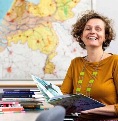 Gezellige vrouw leest kleurrijk kinderboek in klaslokaal met stapel kinderboeken op tafel, achtergrond met overzichtskaart Nederland, passend bij educatie, lezen en scholing, logo RTV SLOS.