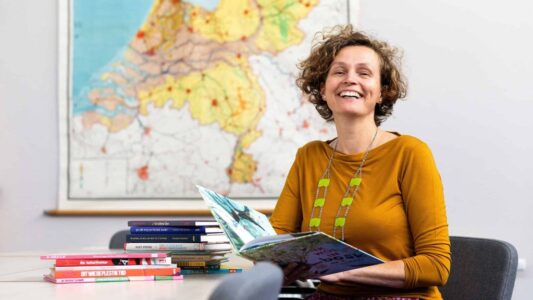 Gezellige vrouw leest kleurrijk kinderboek in klaslokaal met stapel kinderboeken op tafel, achtergrond met overzichtskaart Nederland, passend bij educatie, lezen en scholing, logo RTV SLOS.