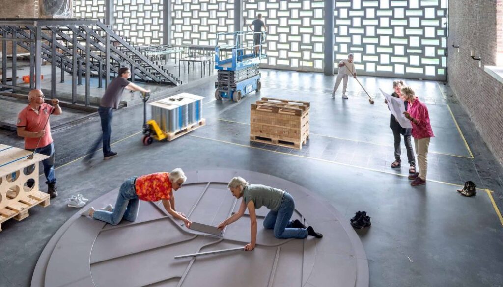 Een groep mensen werkt samen aan het bouwen of installeren van een groot rond object in een industriële ruimte, met verschillende arbeidsactiviteiten en materialen zichtbaar.