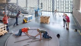 Een groep mensen werkt samen aan het bouwen of installeren van een groot rond object in een industriële ruimte, met verschillende arbeidsactiviteiten en materialen zichtbaar.