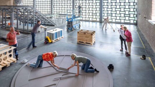 Een groep mensen werkt samen aan het bouwen of installeren van een groot rond object in een industriële ruimte, met verschillende arbeidsactiviteiten en materialen zichtbaar.
