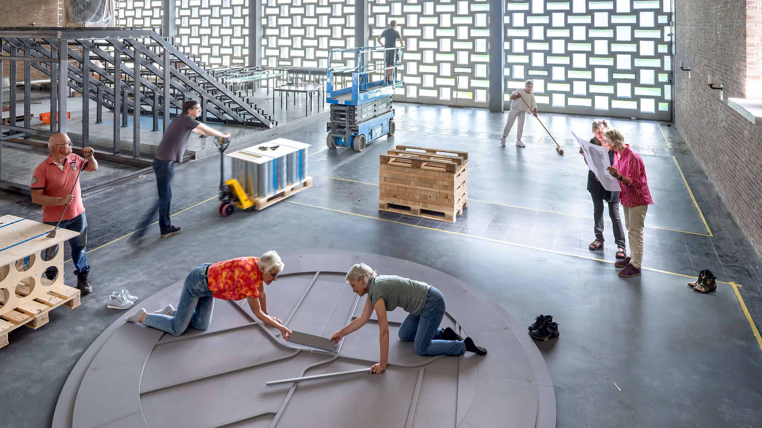 Een groep mensen werkt samen aan het bouwen of installeren van een groot rond object in een industriële ruimte, met verschillende arbeidsactiviteiten en materialen zichtbaar.