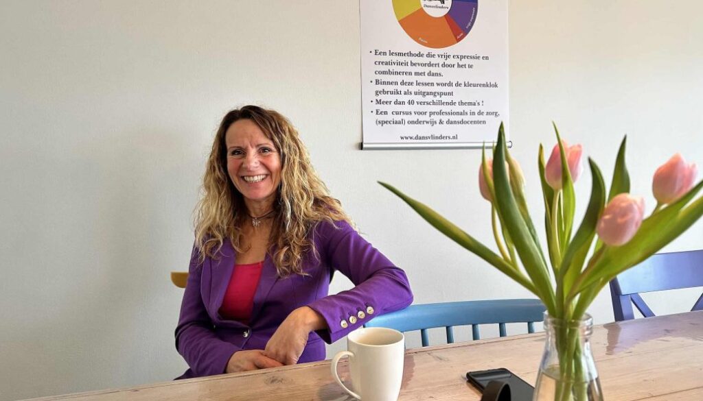 Blije vrouw in een purple blazer zit aan een tafel met een kop koffie en een smartphone, met een vaas met roze tulpen op de voorgrond en een poster over danslessen op de muur achter haar, in een lichte en frisse kantoorruimte.