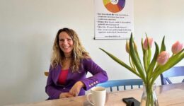 Blije vrouw in een purple blazer zit aan een tafel met een kop koffie en een smartphone, met een vaas met roze tulpen op de voorgrond en een poster over danslessen op de muur achter haar, in een lichte en frisse kantoorruimte.