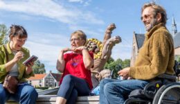 Vrolijke mensen in gesprek op een plein met beelden en een kerk op de achtergrond, glimlachend en genietend van een zonnige dag, benadrukt inclusieve gemeenschap en maatschappelijke betrokkenheid.