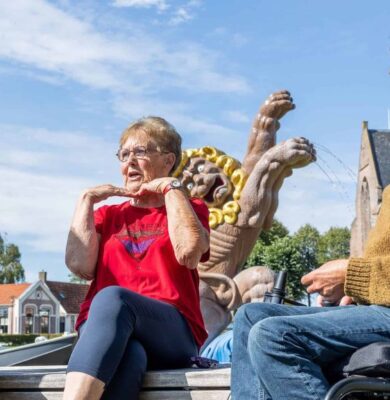 Vrolijke mensen in gesprek op een plein met beelden en een kerk op de achtergrond, glimlachend en genietend van een zonnige dag, benadrukt inclusieve gemeenschap en maatschappelijke betrokkenheid.