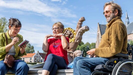 Vrolijke mensen in gesprek op een plein met beelden en een kerk op de achtergrond, glimlachend en genietend van een zonnige dag, benadrukt inclusieve gemeenschap en maatschappelijke betrokkenheid.