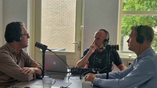 Man die in microfoon spreekt tijdens radio-opname bij RTV SLOS, podcast opname, radiostudio met drie mensen met koptelefoons, gesprek over lokale media en community nieuws in Nederland.