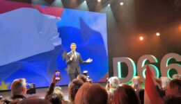Informatieve presentatie op evenement met grote LED-scherm, spreker in pak, publiek met camera's en vlaggen, landelijke sfeer, prominent "D66" logo op achtergrond, podiumverlichting, Politieke bijeenkomst, verkiezingscampagne.