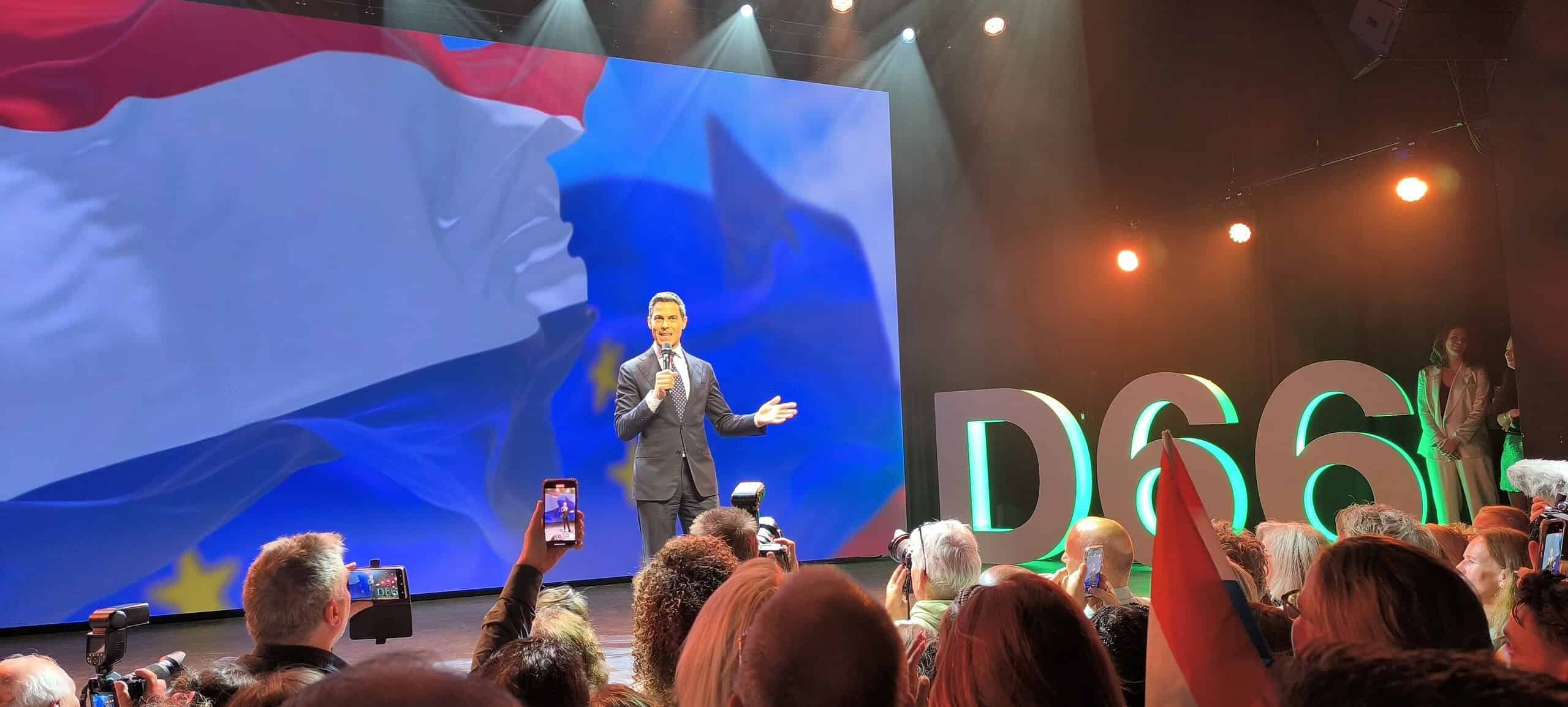 Informatieve presentatie op evenement met grote LED-scherm, spreker in pak, publiek met camera's en vlaggen, landelijke sfeer, prominent "D66" logo op achtergrond, podiumverlichting, Politieke bijeenkomst, verkiezingscampagne.